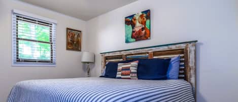 A bedroom with a bed, a window with blinds, a painting on the wall, and a lamp.