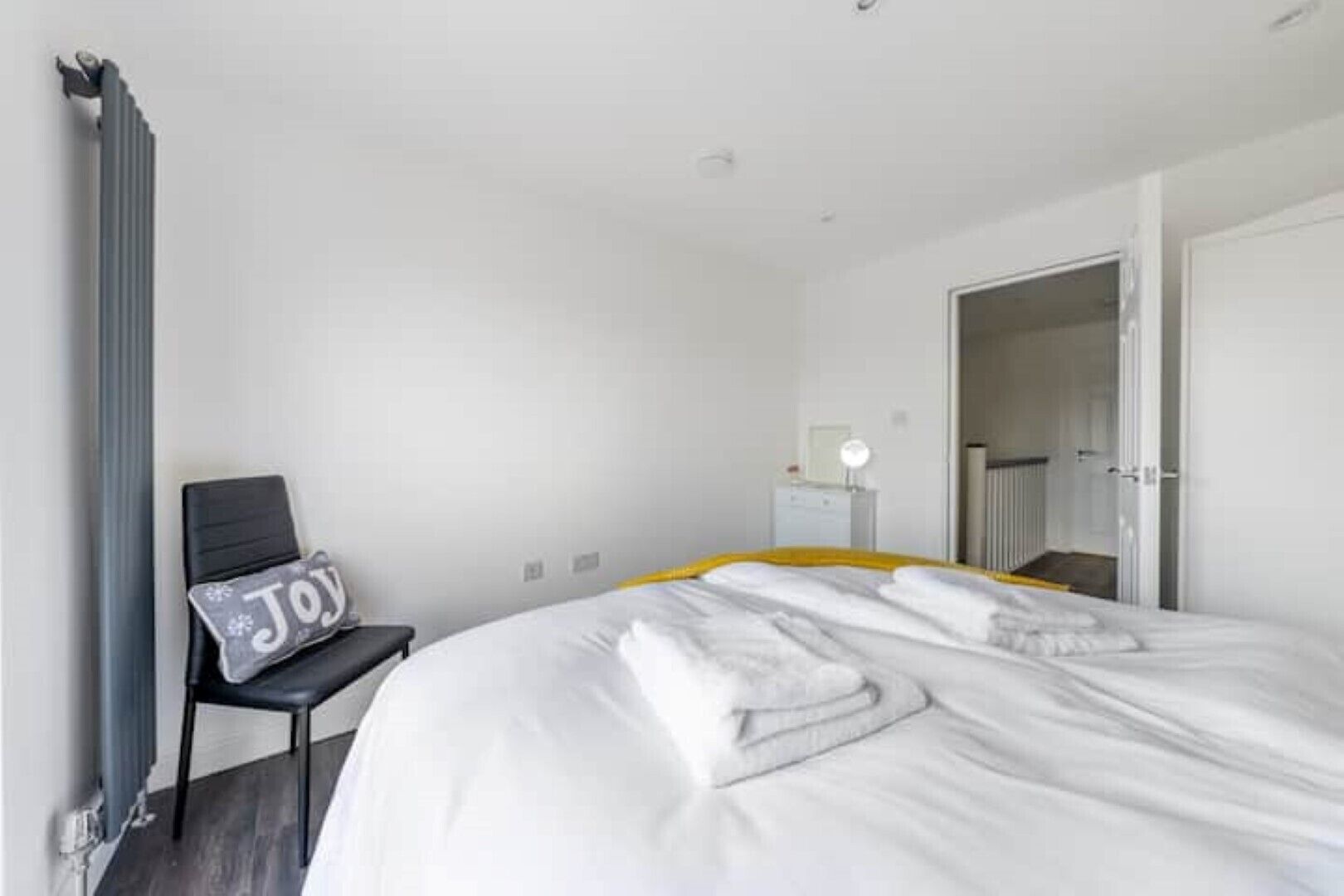 A hotel room with a bed, a chair with a pillow, a bedside table, and a radiator.