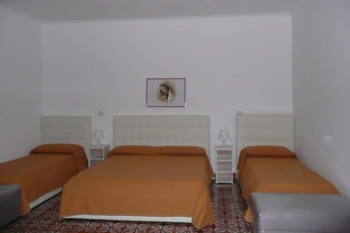 A hotel room with three beds, each with an orange bedspread, white headboards, and bedside tables with lamps. A framed picture hangs on the wall above the beds.