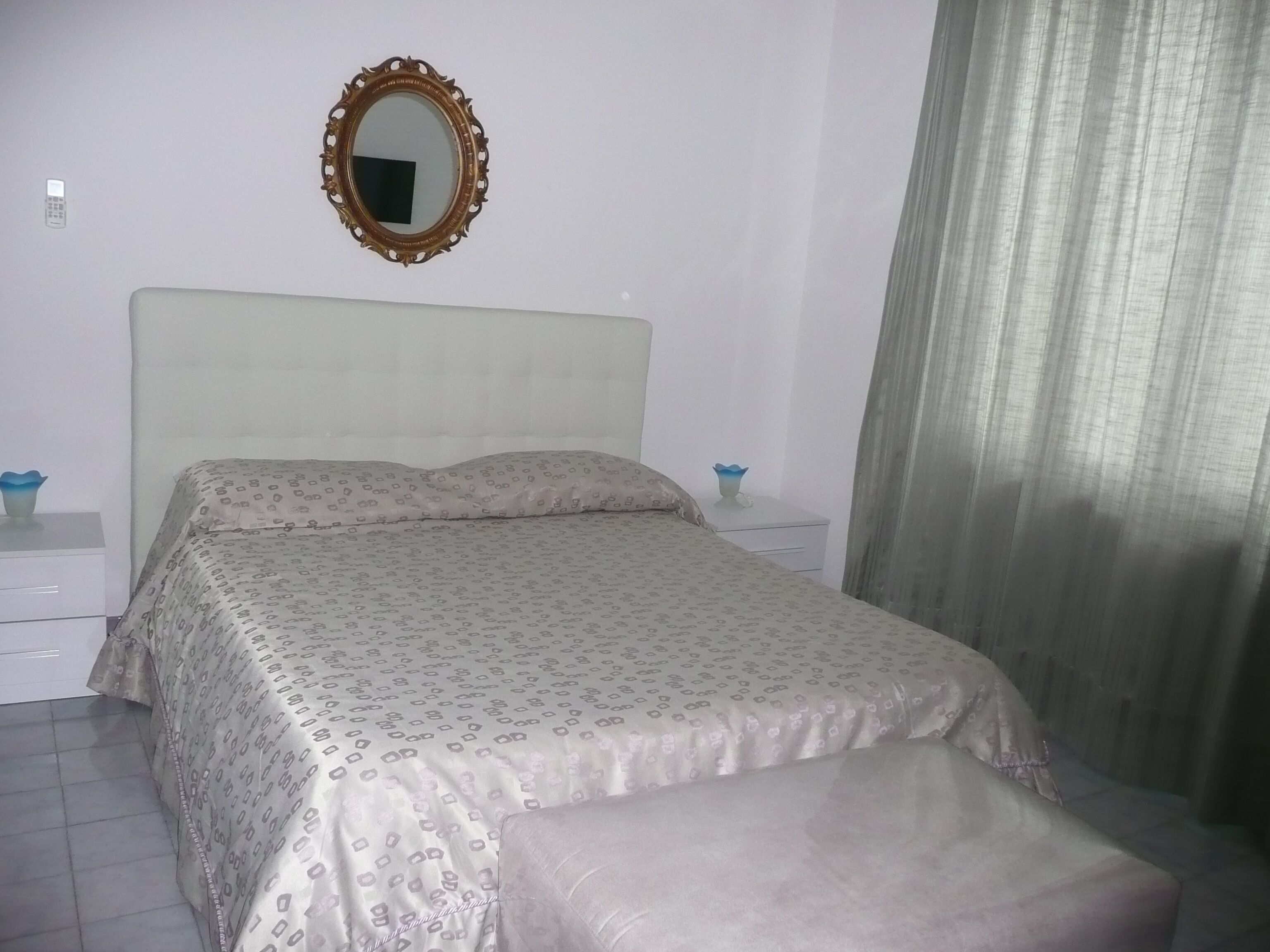 Un dormitorio con cama, mesita de noche, espejo y ventana con cortinas.