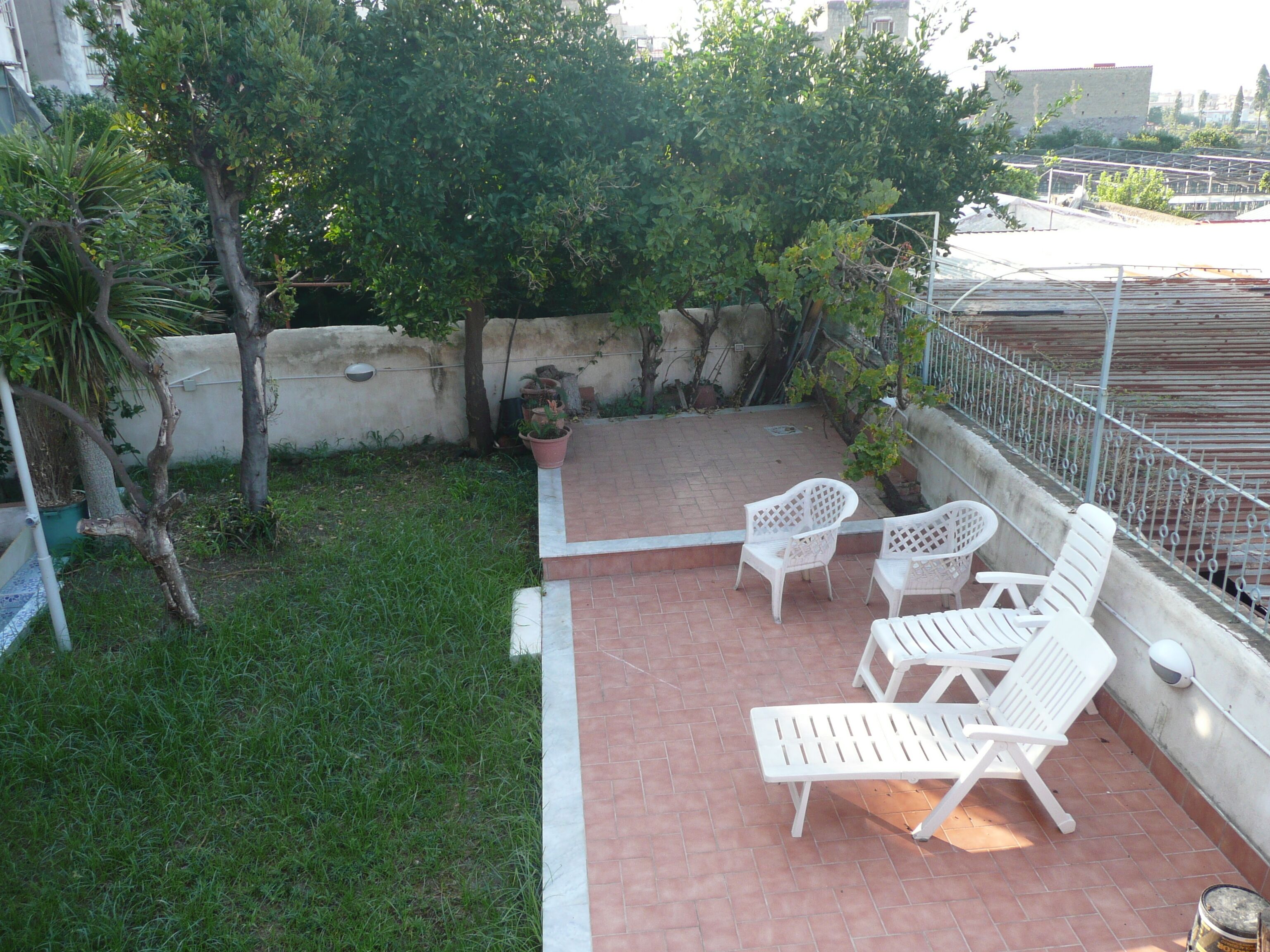 A patio area with white plastic chairs and a small table, surrounded by a low brick wall and greenery.
