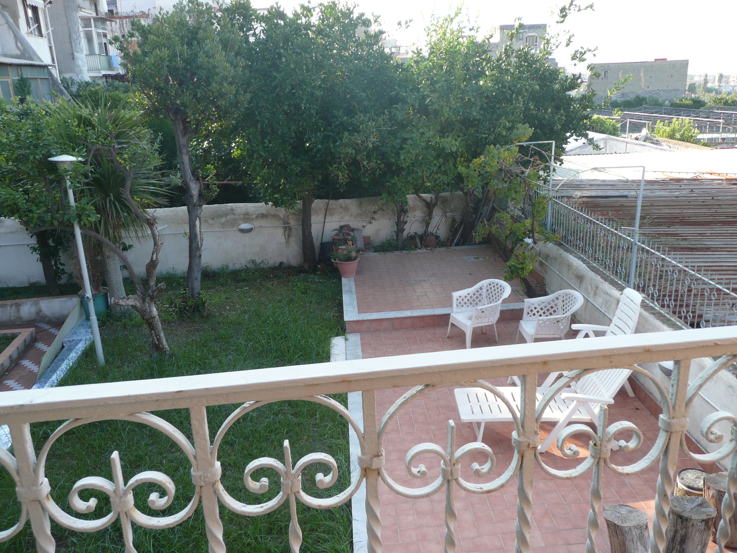 A balcony with a wrought iron railing, a small patio area with white plastic chairs, and a view of a garden with trees and a wall.