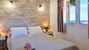 A bedroom with a stone wall, a bed with white bedding, a nightstand with a lamp, and a window with curtains. - Corsica seaside House n°5 Residence Saint Vincent Corse (Occhiatana)