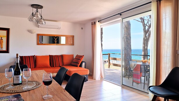 A room with a wooden dining table, a sofa with orange cushions, a bottle of wine, and a glass of red wine. There is a large window with a view of the ocean and a tree. - Corsica seaside House n°5 Residence Saint Vincent Corse (Occhiatana)