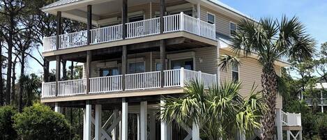 A two-story beachfront house with a balcony on each floor, supported by wooden stilts.