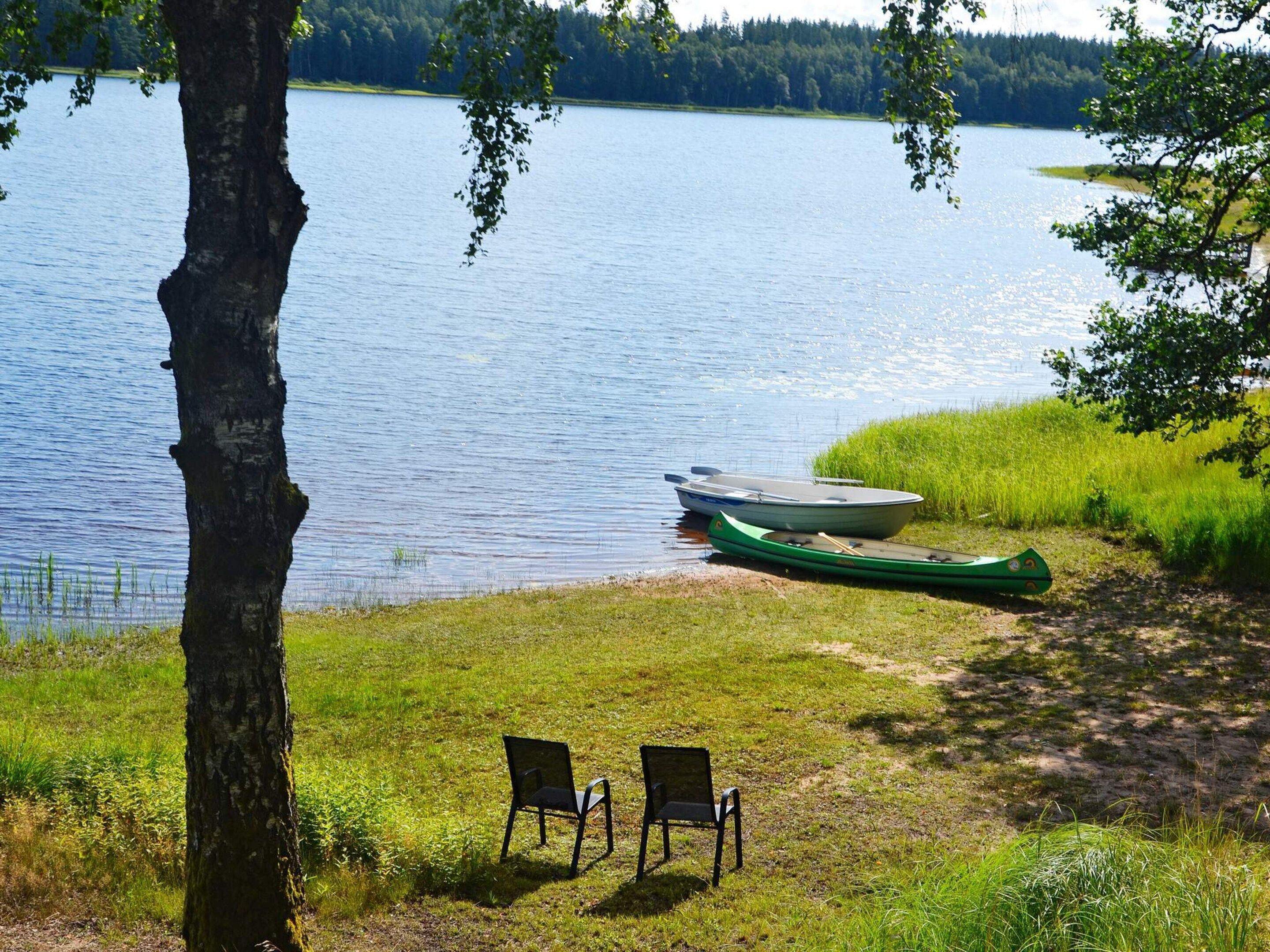 A lakeside view with a boat docked, two chairs on the grass, and a tree in the foreground.