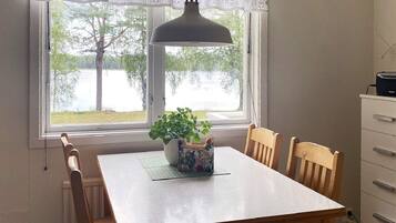 A dining area with a table, chairs, a potted plant, a window with a view of trees and a body of water, and a wall-mounted lamp.