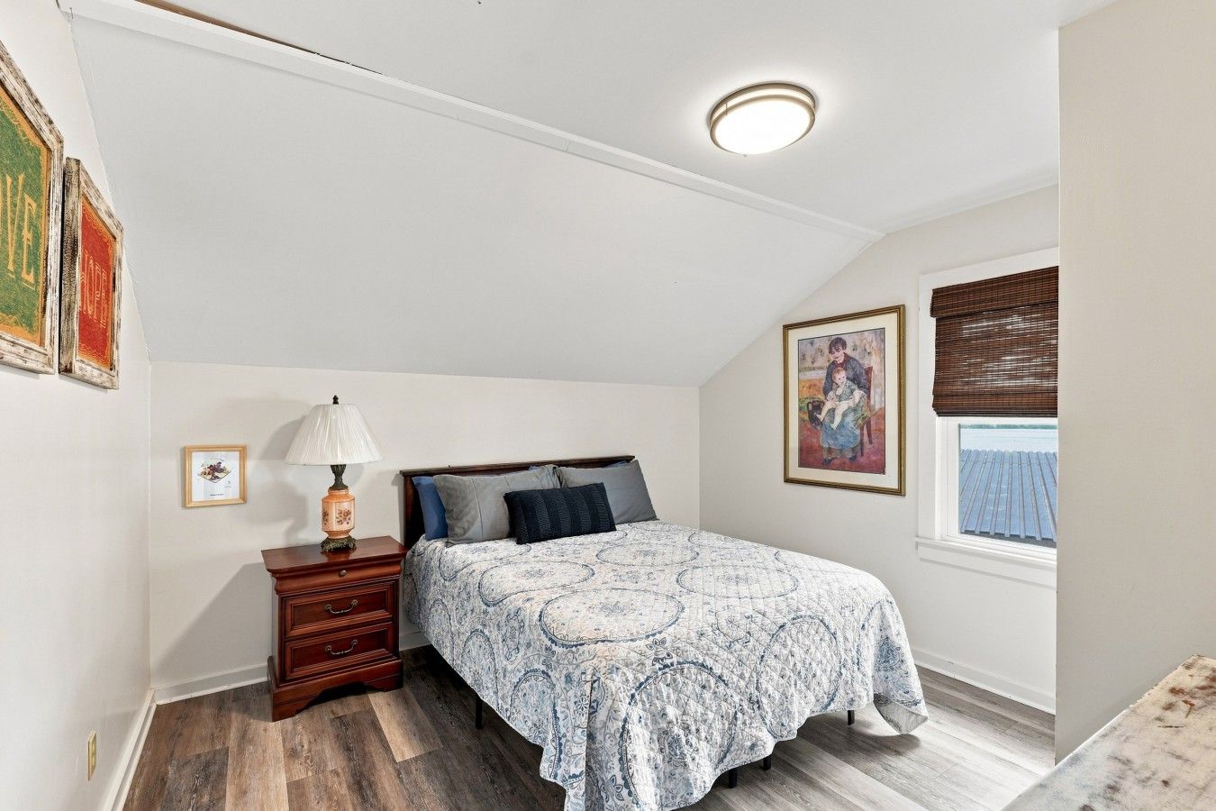 A bedroom with a bed, a nightstand, a lamp, a window with blinds, and artwork on the walls.