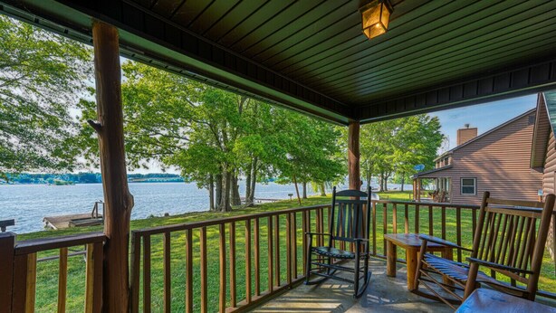 A covered deck with a view of a lake, wooden furniture, and a house in the background.