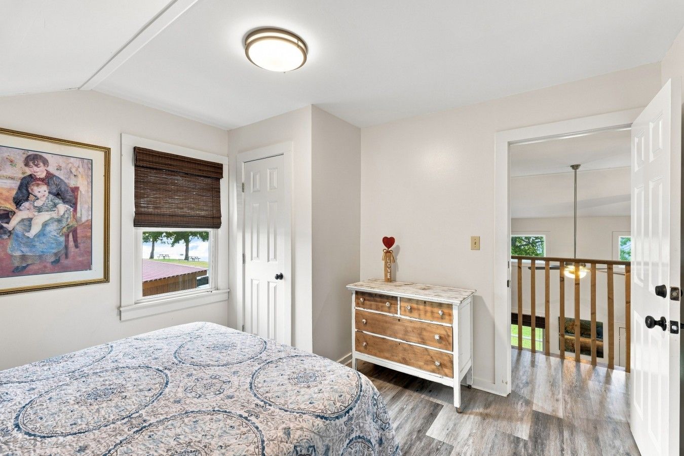 Un dormitorio con cama, cómoda, un cuadro, ventana con persianas y puerta que da a un balcón.
