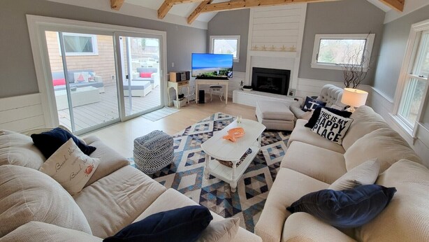 A spacious living room with a high ceiling, a fireplace, a television, and a sliding glass door leading to an outdoor area. - Sprawling 5 BR Chatham Escape / Endless Outdoor Living & Entertaining Choices (Chatham)