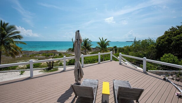 A wooden deck with two chairs and a table, overlooking a beach with turquoise water and palm trees. - Newly Remodeled Home on Calm Caribbean w/Sunset Views, Snorkeling, Generator! (N Palmetto Point)