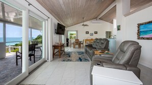 A spacious living area with a large window overlooking the ocean, a ceiling fan, and a modern kitchen in the background. - Newly Remodeled Home on Calm Caribbean w/Sunset Views, Snorkeling, Generator! (N Palmetto Point)