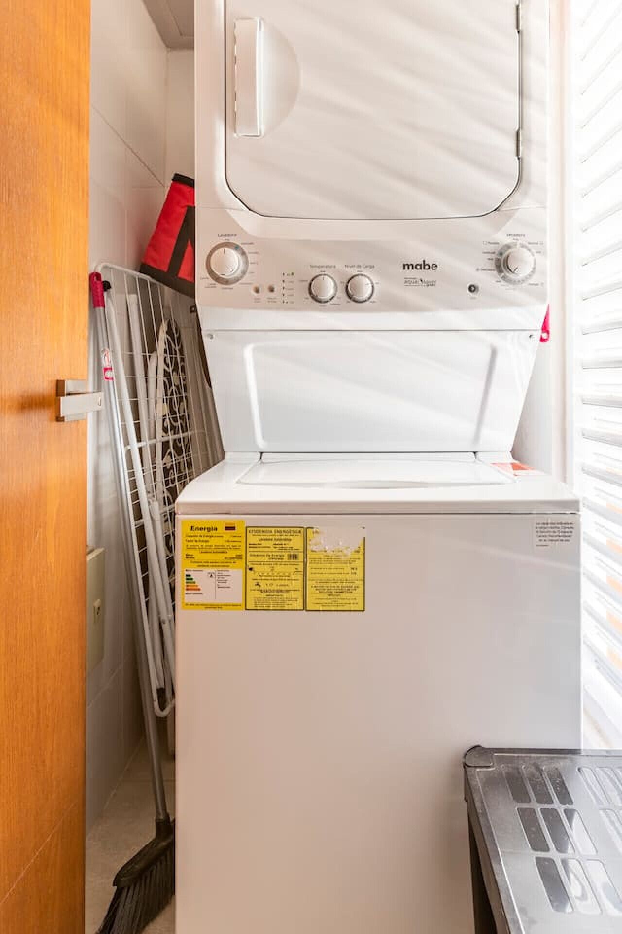 A compact, white washing machine with a control panel, situated in a narrow laundry room with a wooden door and a metal rack.