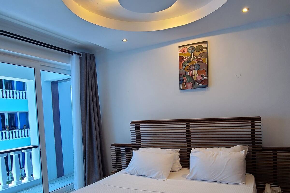 A modern hotel room with a large bed, a ceiling light fixture, and a painting on the wall.