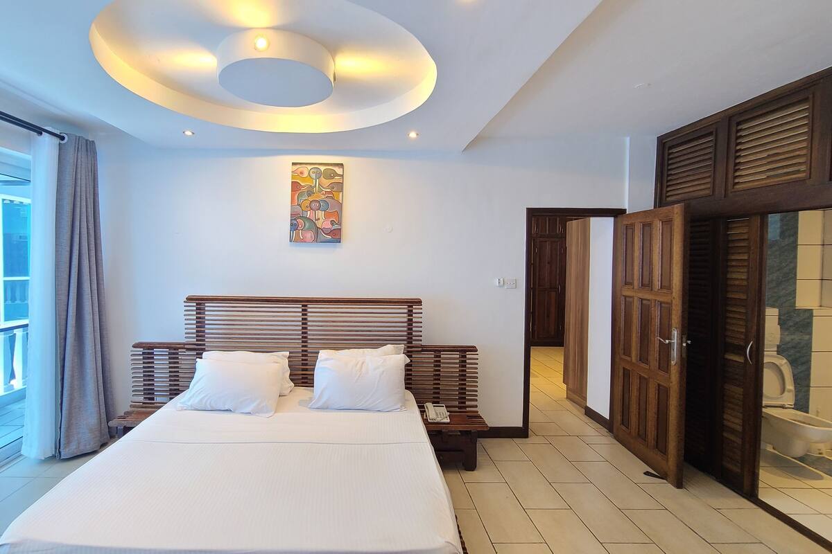 A modern hotel room with a large bed, a ceiling light fixture, a painting on the wall, and a view of the outside.