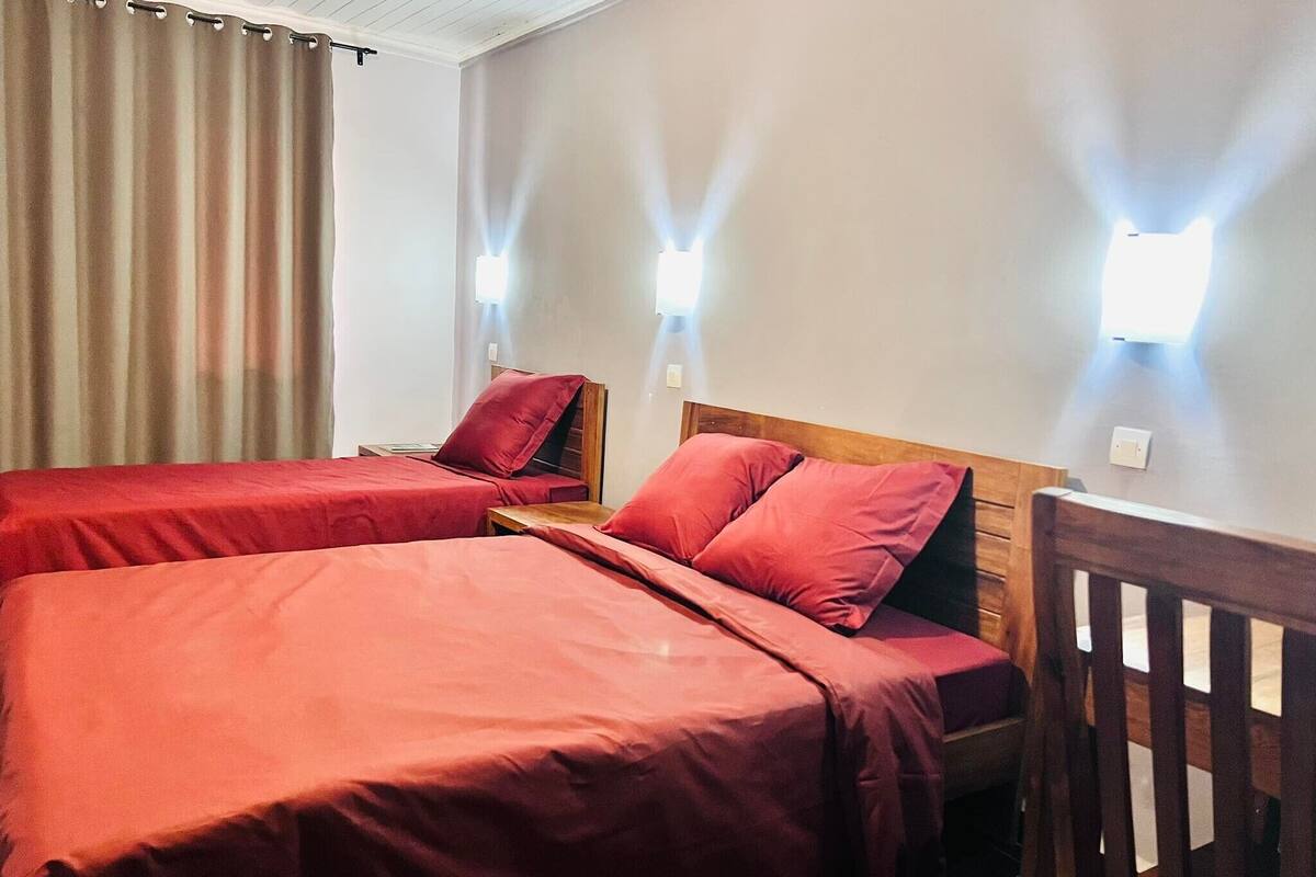 A hotel room with two beds, each with red bedding, wooden headboards, and bedside tables. There are two wall-mounted lights and a window with curtains.