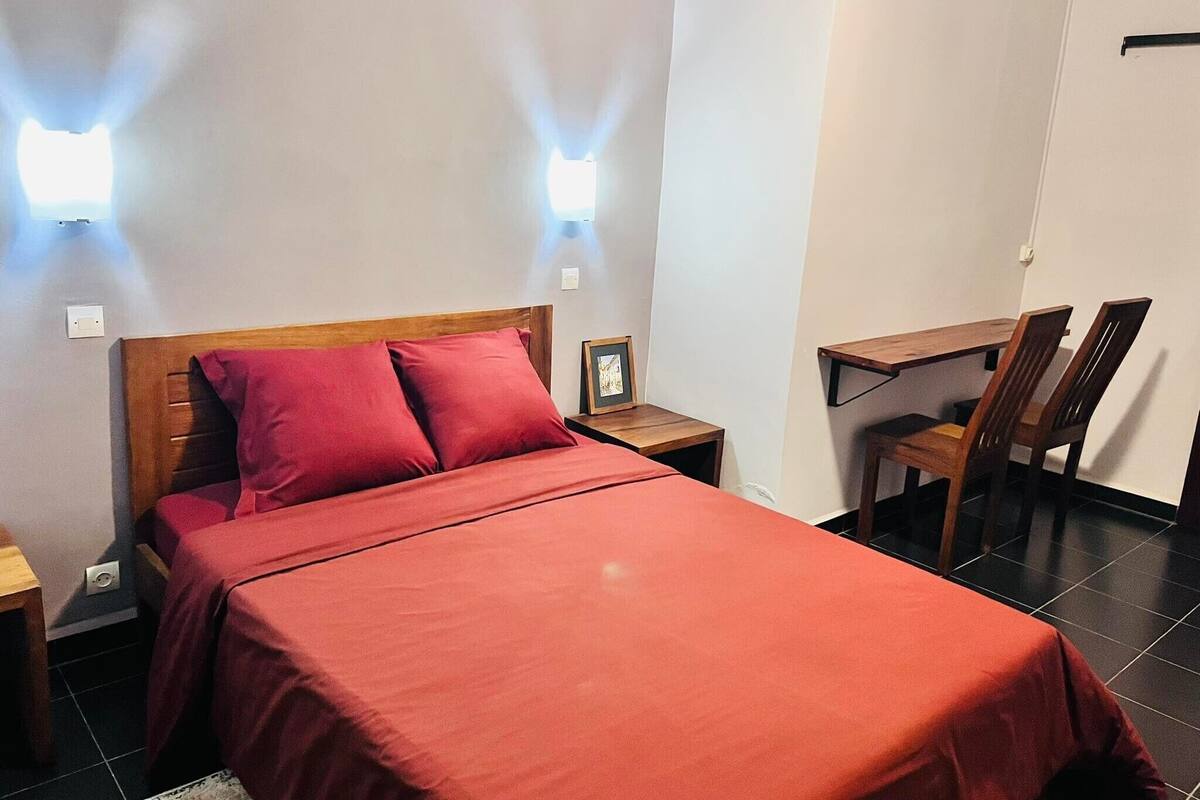 A neatly made bed with a red bedspread and pillows, a wooden nightstand with a framed picture, and a desk with two chairs in a room with light-colored walls and tiled flooring.