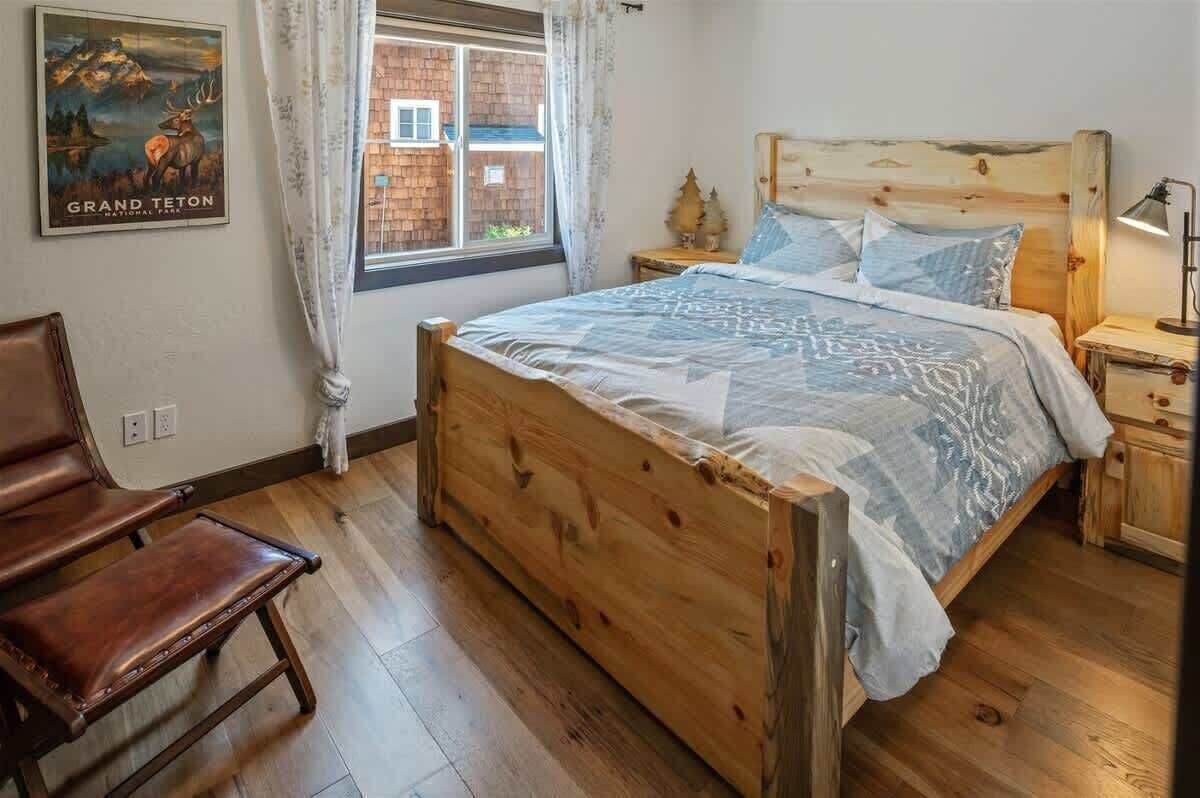 A wooden bed with a patterned bedspread, a nightstand with a lamp, a chair, and a window with curtains.