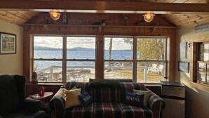 A cozy cabin with a plaid sofa and armchairs, large windows overlooking a lake, and a wooden ceiling.