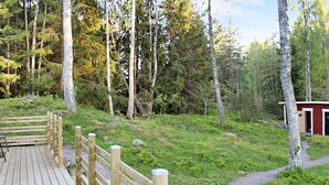 A wooden deck with a railing, surrounded by a forested area with a small building in the background. - 7 Person Holiday Home in Skyllberg (Skyllberg)