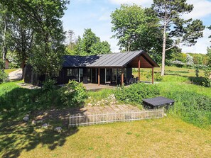 A modern cabin with a covered patio, surrounded by lush greenery and a wooden fence. - 5 Star Holiday Home in Millinge (Millinge)