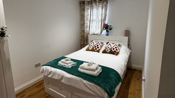 A neatly made bed with a green throw, white bedding, and decorative pillows. There is a small window with curtains, a ceiling light, and a wall-mounted outlet. - cosy and clean room- 2 minuets away from train station (London)