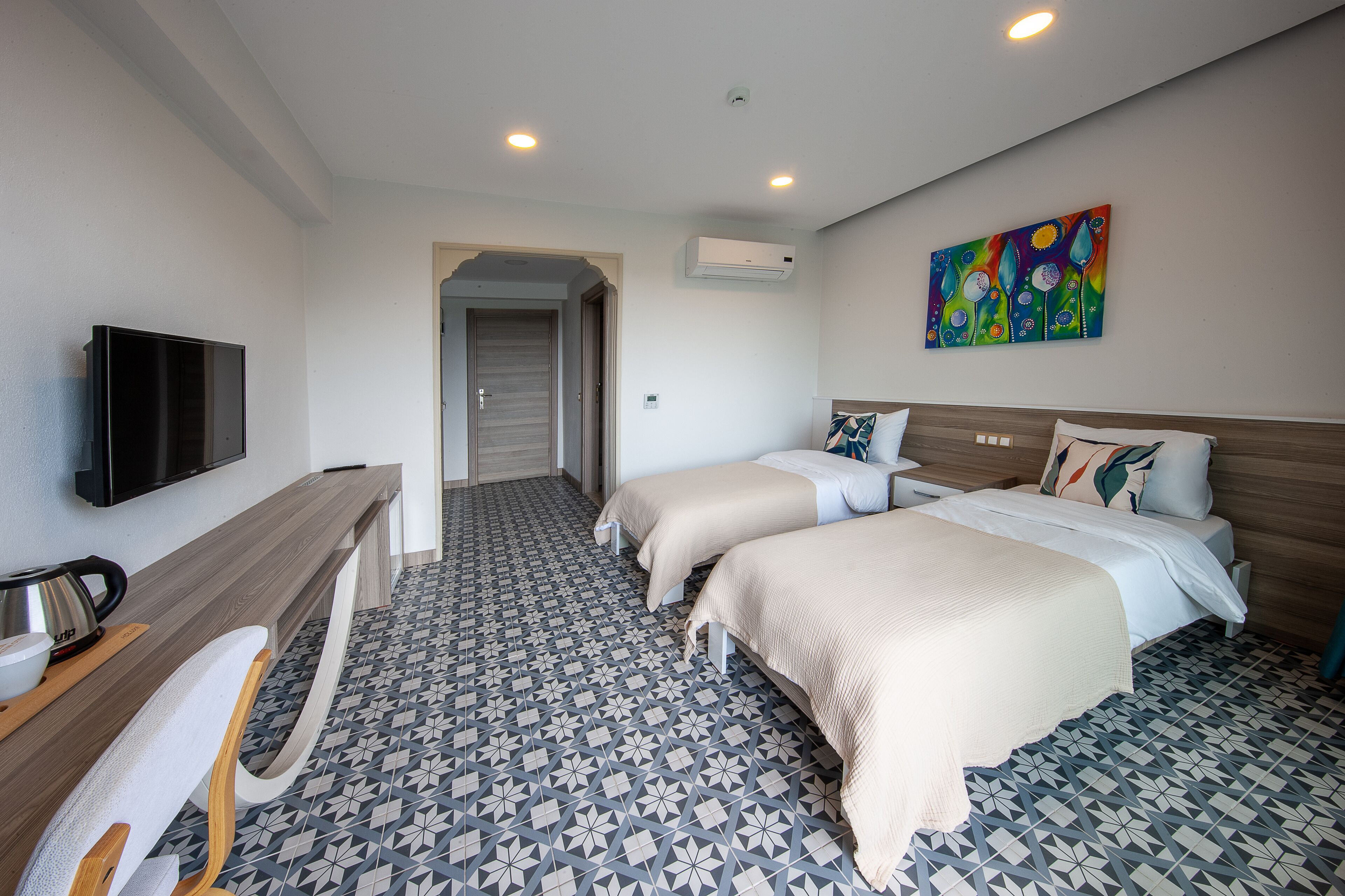 A hotel room with two beds, a TV, a desk with a kettle, and a colorful painting on the wall.