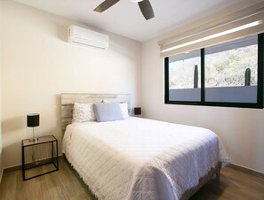 A bedroom with a bed, bedside tables, an air conditioning unit, a ceiling fan, and a window with a view. - Sea Esta is a ground-floor 2-BD, 2-BR condo within walking distance of downtown Cabo, the marina, and Cannery Beach. Featuring an open-concept living space, private balcony, and views of the city and the Sea of Cortez, it’s ideal for a relaxing Cabo. (Cabo San Lucas)