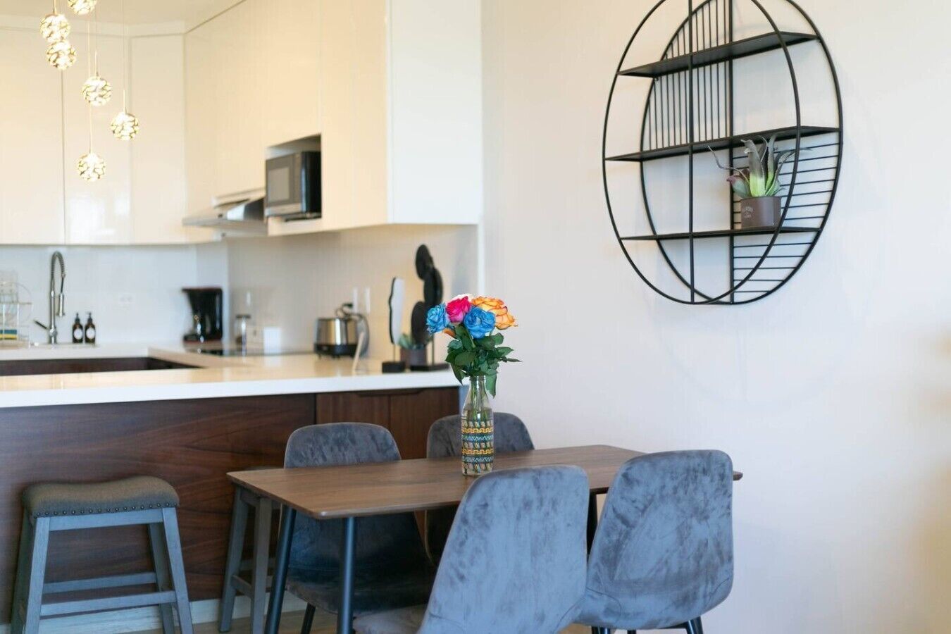 A modern kitchen with a white countertop, a microwave, and a coffee machine. There is a dining table with chairs and a vase with flowers.