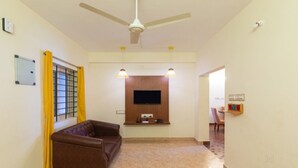 Una sala de estar con un sofá marrón, un ventilador de techo, un televisor de pantalla plana montado en la pared y una pequeña mesa con un mueble para televisor.