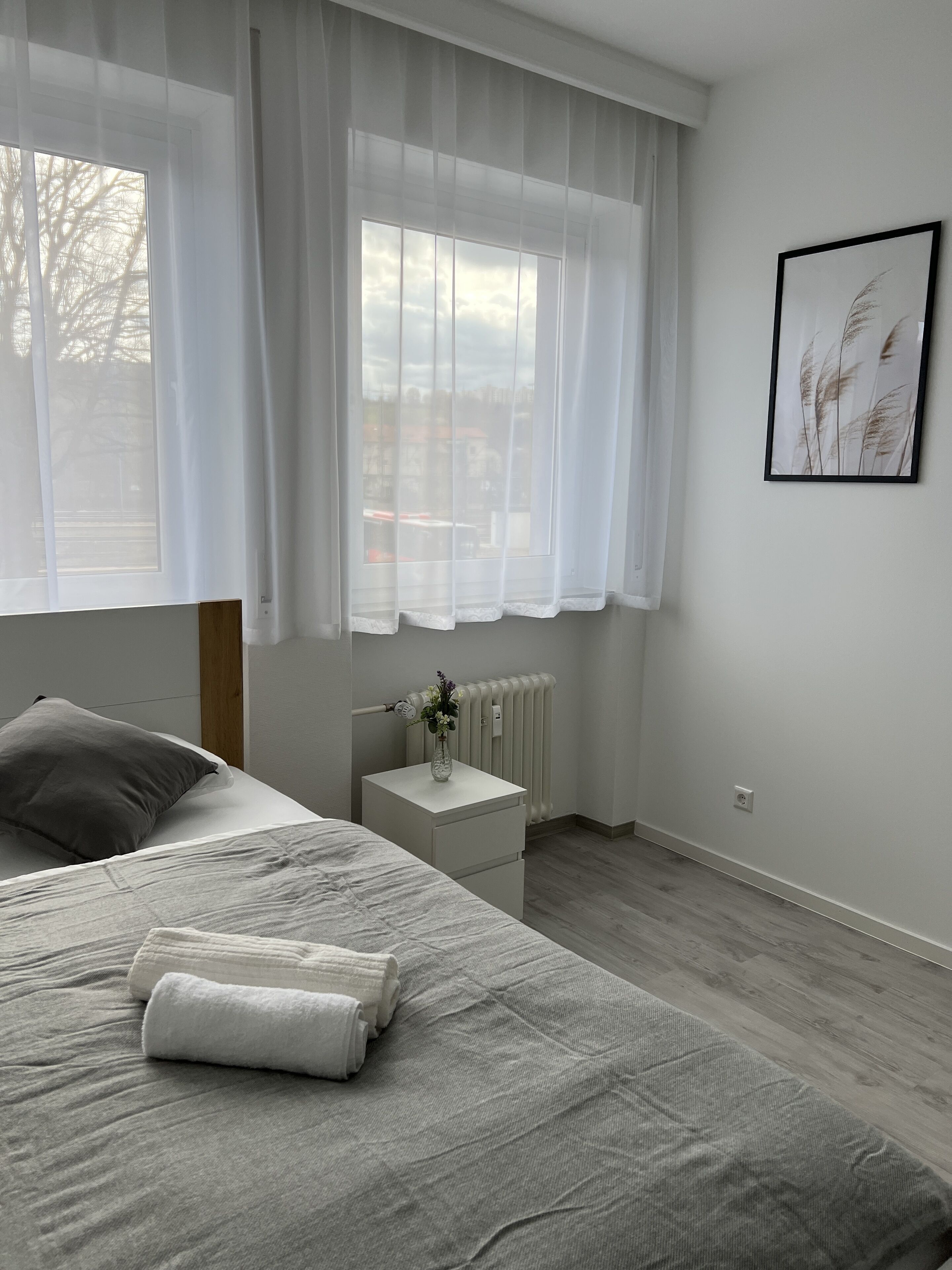 A bedroom with a bed, a nightstand with a vase, a radiator, and a window with sheer curtains.