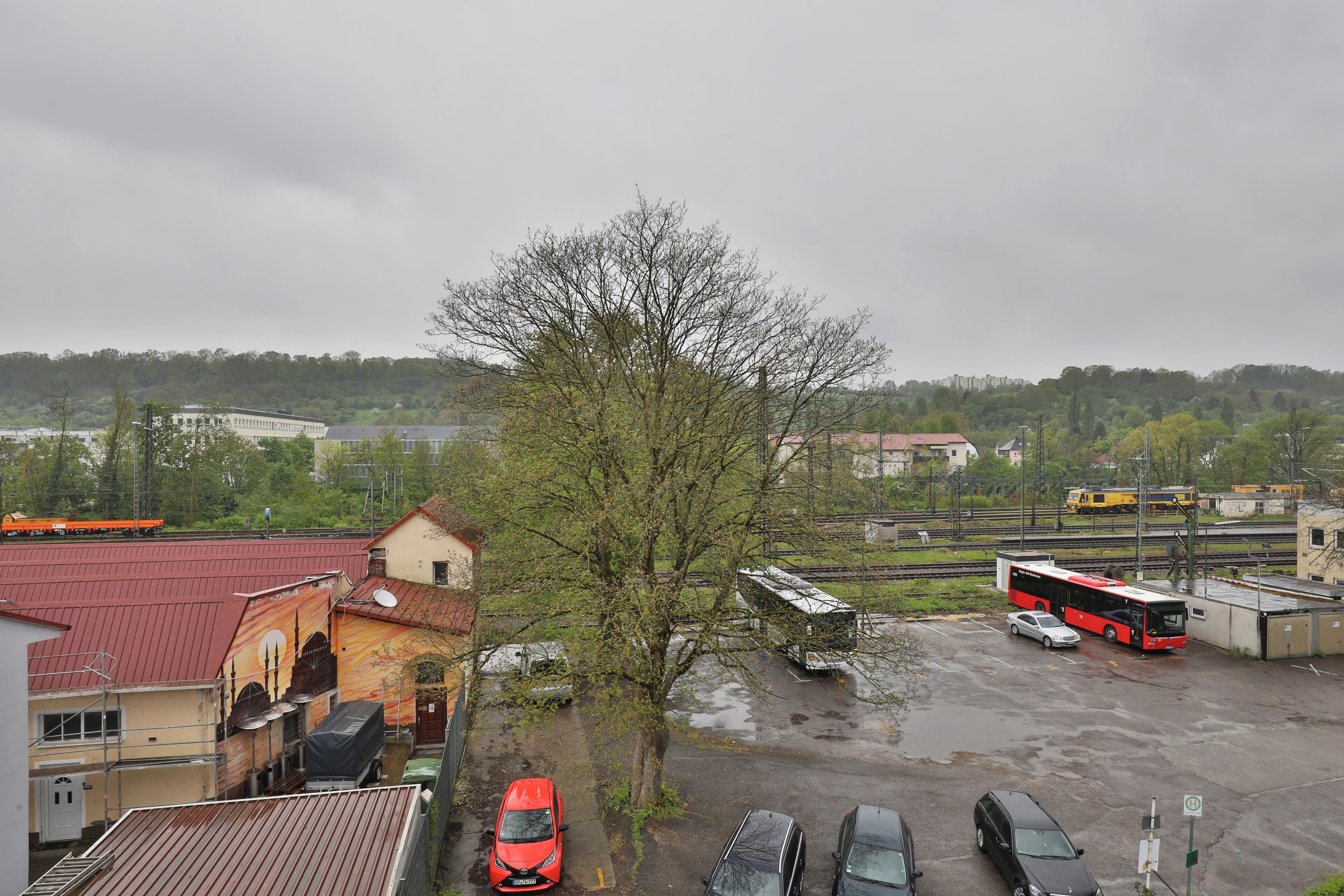 Um estacionamento com vários veículos, incluindo um carro vermelho, um carro branco e um ônibus vermelho. Há edifícios com telhados vermelhos e uma árvore ao primeiro plano.