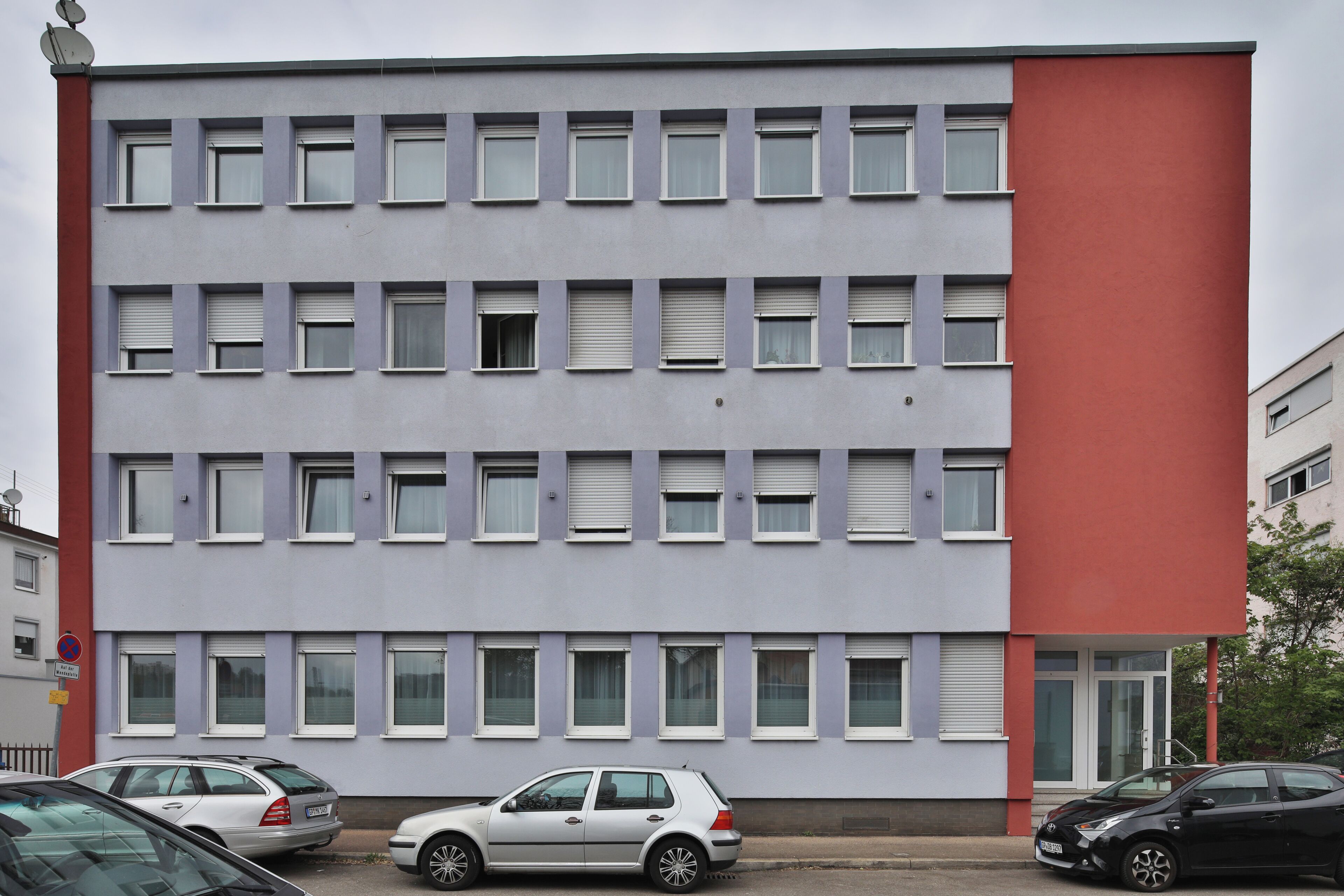 A multi-story building with a red and grey facade, several windows, and parked cars in front.