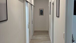 Corredor com piso de madeira, paredes brancas e quadros emoldurados. Há portas em ambos os lados e um lustre no teto.