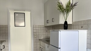 Uma cozinha moderna com armários brancos, lava-louças embutida e frigorífico branco. Há uma pequena planta na bancada e um painel de azulejos atrás do fogão.