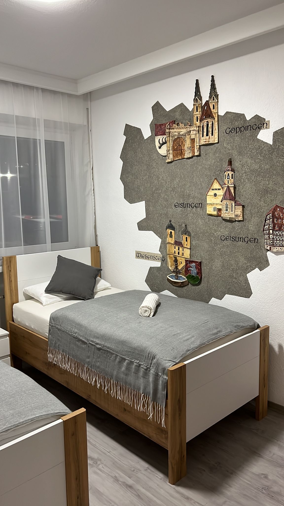 A modern bedroom with a wooden bed, a grey blanket, and a wall map featuring town names and buildings.