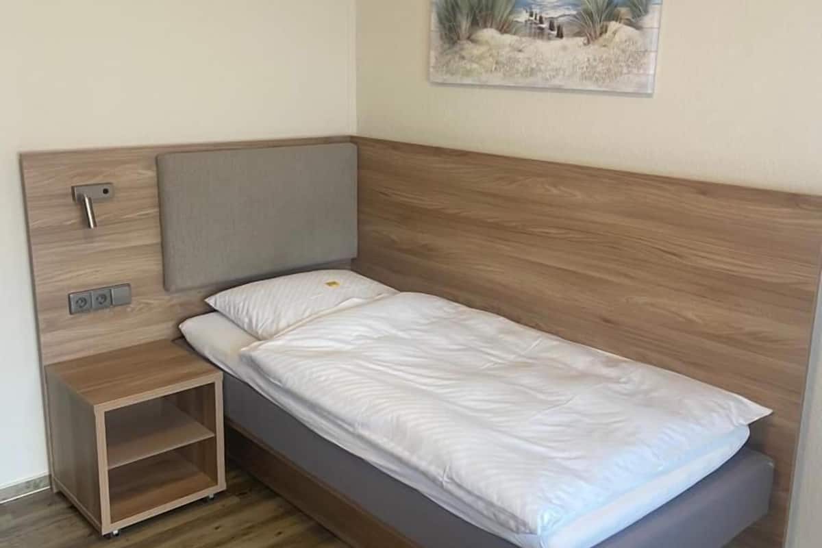 Una habitación pequeña con cama individual, cabecera de madera, mesita de noche y un cuadro colgado en la pared.