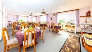 A dining area with multiple round tables, each with a purple tablecloth and wooden chairs. There are ceiling fans and a coffee station with a sign. - Hotel am Stern (Uelzen)