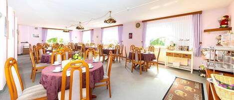 A dining area with multiple round tables, each with a purple tablecloth and wooden chairs. There are ceiling fans and a coffee station with a sign.