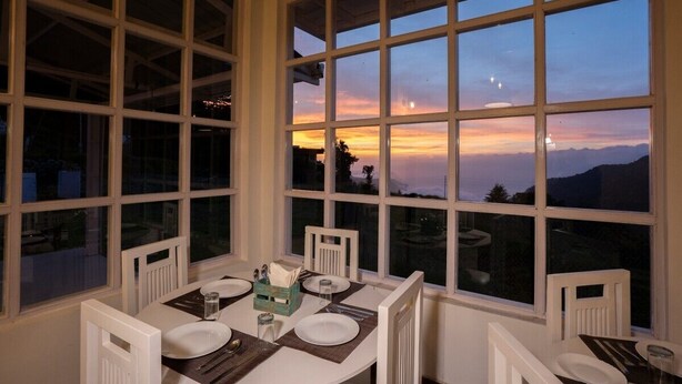 Un coin repas avec une table dressée pour deux personnes, de grandes fenêtres offrant une vue sur le coucher de soleil et les montagnes.