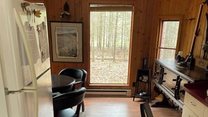 A cabin interior with a dining table, chairs, a refrigerator, a vacuum cleaner, and a window with a forest view.