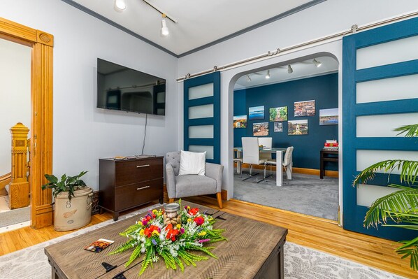 Ein modernes Hotelzimmer mit einem Flachbildfernseher, einem grauen Sessel, einem Holztisch mit einer Blumenkomposition und einer Schiebetür zu einem Essbereich mit Blick auf die Stadt.