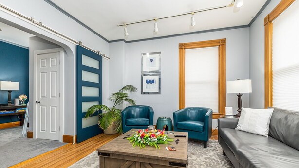 A living room with a wooden coffee table, two blue armchairs, a grey sofa, and a large window with blinds. - The Dubuque House - Historic Downtown Location (Dubuque)