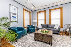 Ein Wohnzimmer mit blauem Sofa, grauem Sofa, Couchtisch aus Holz und einem großen Fenster mit Jalousien.