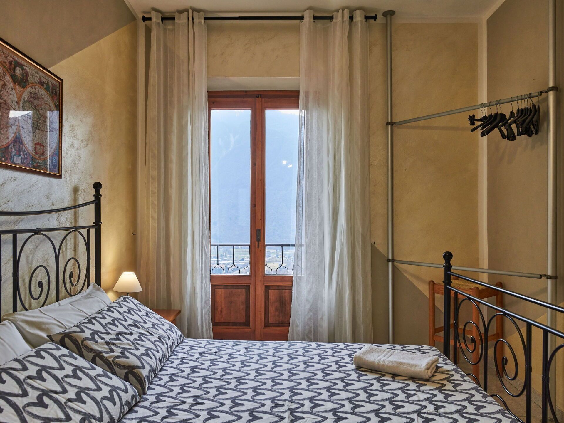A bedroom with a bed, a window with curtains, a map on the wall, and a metal rack with shoes.