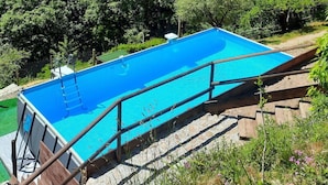 En swimmingpool med ett trädäck och en stege, omgiven av frodig grönska och med ett bergigt landskap i bakgrunden.