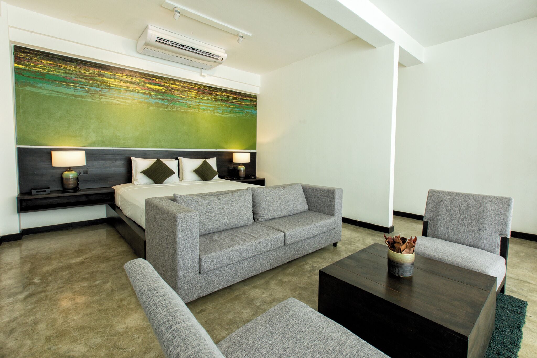 Ein modernes Hotelzimmer mit grauem Sofa, einem kleinen Sessel, einem Couchtisch und einem Bett mit Kopfteil. An der Wand hängt ein großes abstraktes Gemälde und über dem Bett befindet sich eine Klimaanlage.