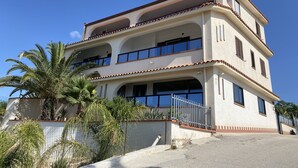 A two-story building with balconies, a gated entrance, and surrounding palm trees. - Nora's Flats - Trilo Apartament (Triscina)