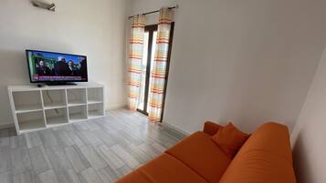 Een woonkamer met een witte tv-meubel, een oranje bank en een televisie waarop een nieuwsuitzending draait.
